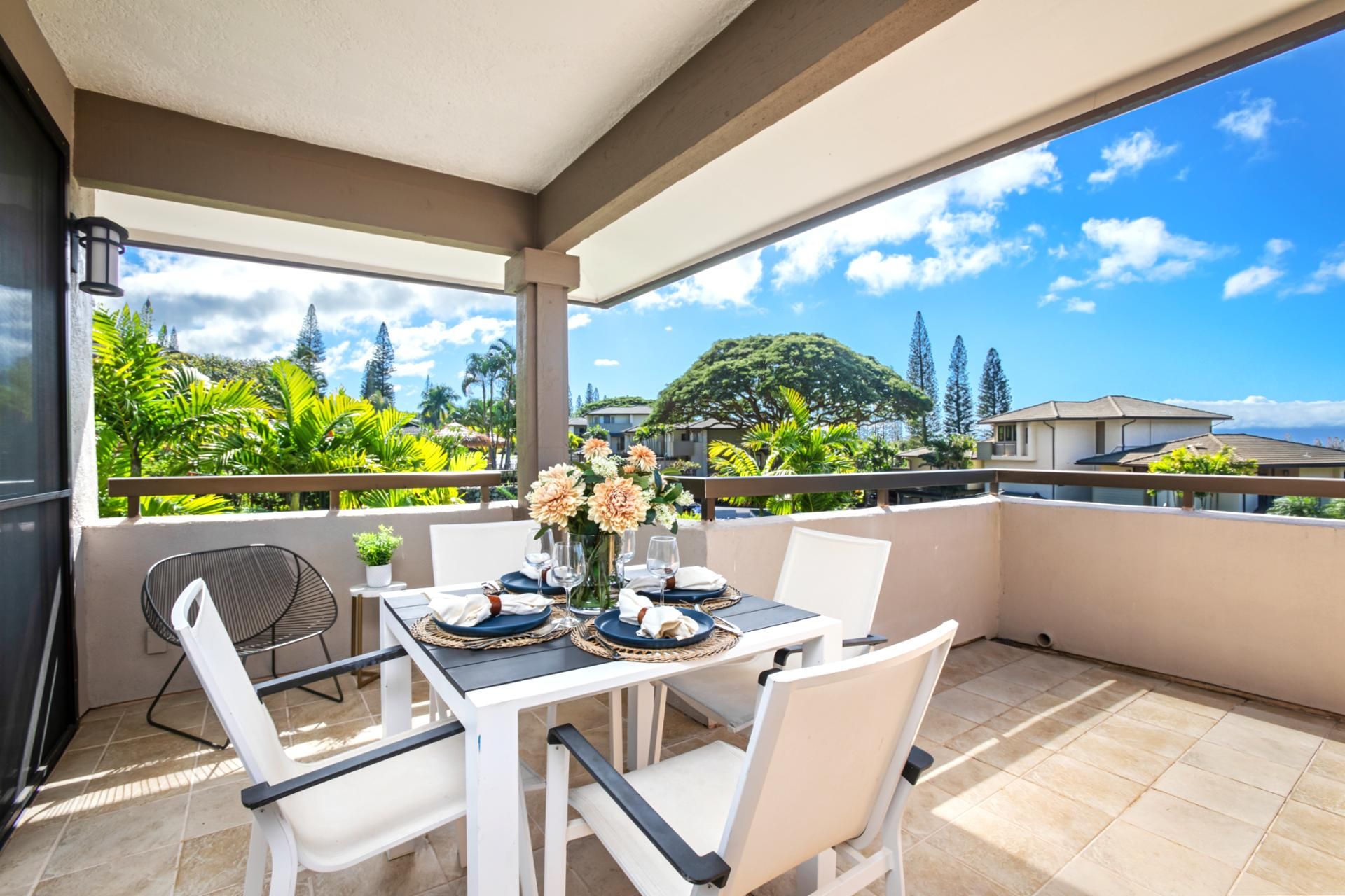 A covered outdoor dining area with a table set for two, surrounded by white chairs, and overlooking a lush landscape with trees and houses.