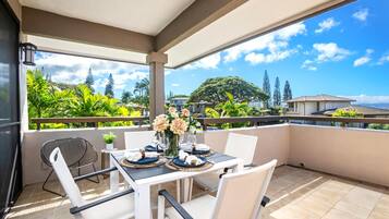 A covered outdoor dining area with a table set for two, surrounded by white chairs, and overlooking a lush landscape with trees and houses.