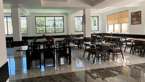 A spacious dining area with multiple tables and chairs, large windows offering views of greenery, and a polished marble floor. - Hotel Choskor (Leh)