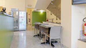 Angolo cottura con frigorifero, microonde e macchina del caffè. Sono presenti due sedie e un tavolo con piano in vetro. Un estintore è fissato alla parete.