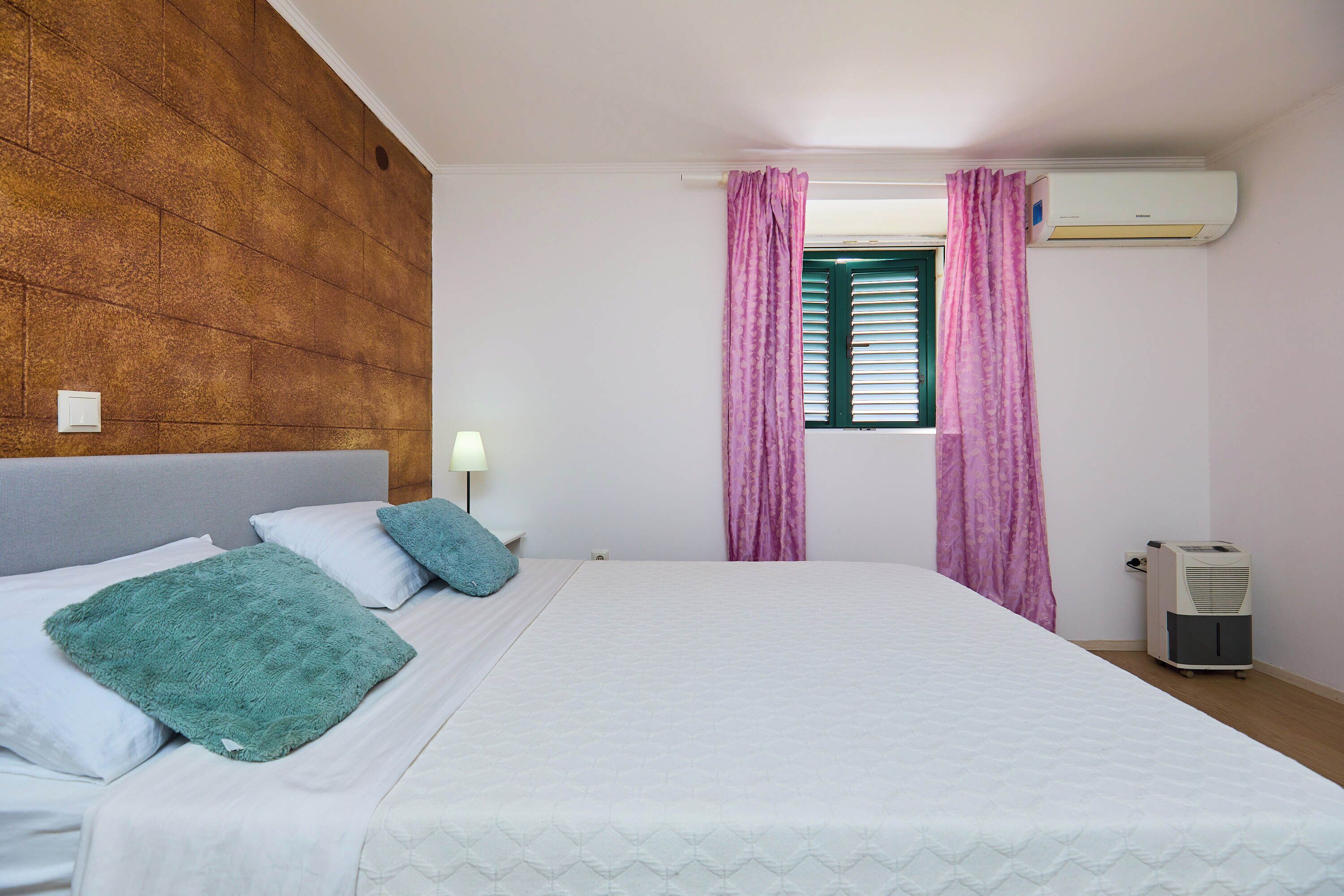 A bedroom with a bed, pillows, a nightstand, a lamp, a window with curtains, and an air conditioning unit.