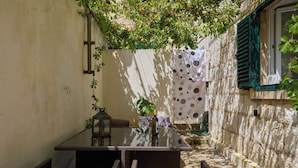 Un patio exterior estrecho con mobiliario de mimbre, una mesa de vidrio y vistas a un edificio con cortinas de lunares.