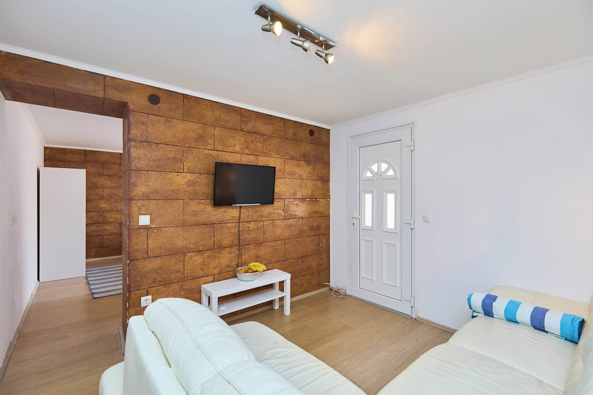 A modern bedroom with a brick accent wall, a flat-screen TV, a small white table with a fruit bowl, a white sofa, and a bed with a striped pillow.