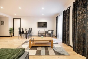 A modern living room with a wooden coffee table, a TV mounted on the wall, a dining area with chairs, and a large window with curtains. - Appartement Confort & Serenite 2bdr/4p (Cannes)