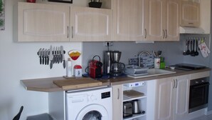A compact kitchen with light wooden cabinets, a built-in washing machine, a coffee maker, and various kitchen utensils.