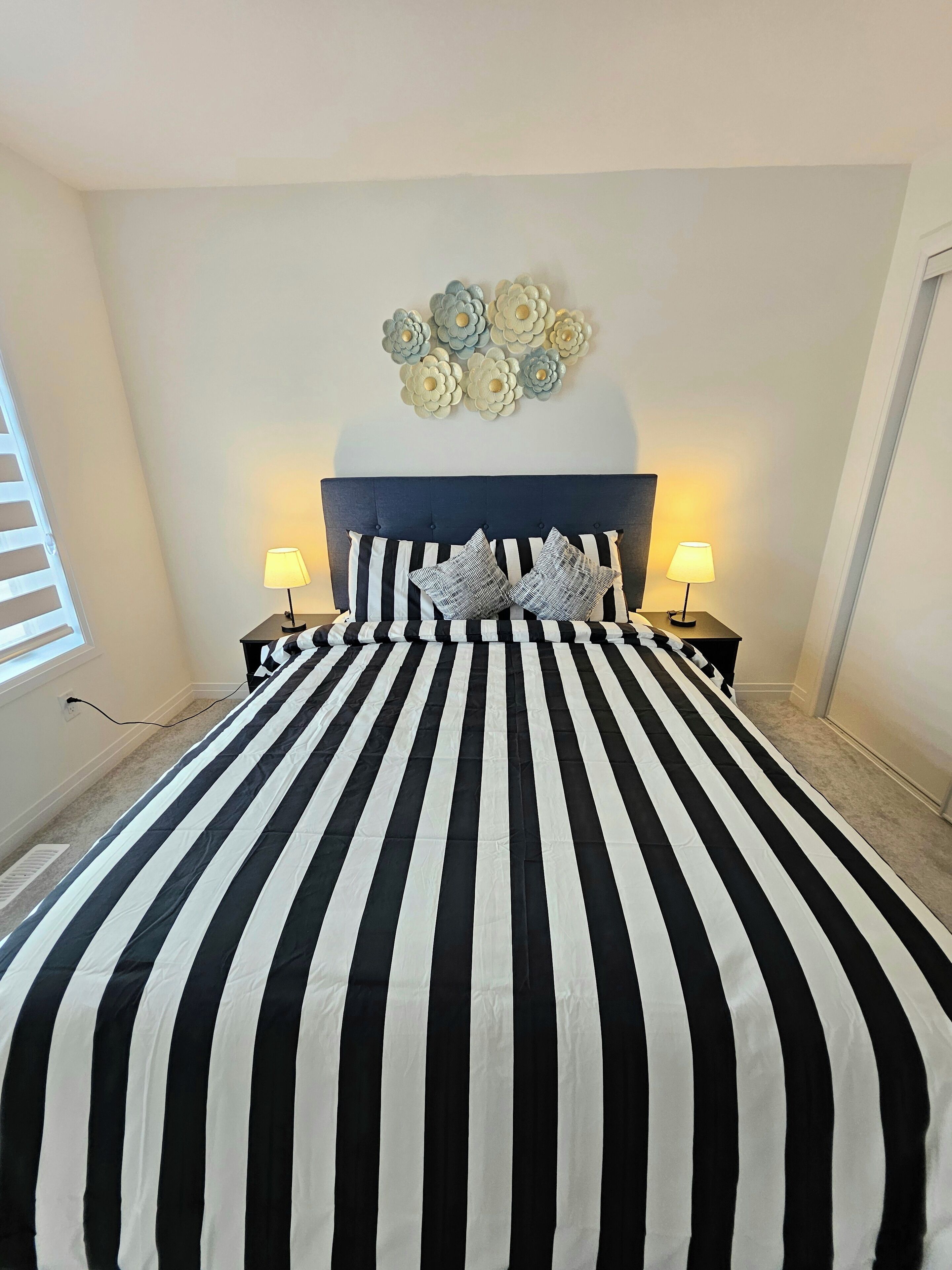 Une chambre avec une couvre-lit à rayures noires et blanches, une tête de lit sombre, deux lampes de chevet et une œuvre d’art murale au-dessus du lit.