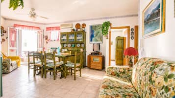 A living room with a sofa, a dining table with chairs, a cabinet with dishes, a television, and a ceiling fan.