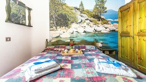 A bed with a colorful quilt, a tray with breakfast items, and a painting of a rocky landscape with a body of water. - Residence 4 Colonne (Bosa)