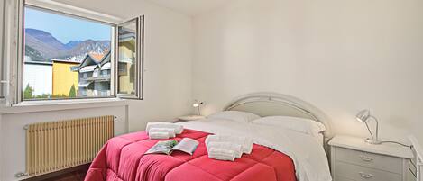 A bedroom with a large window, a bed with red bedding, a bedside table, and a nightstand.