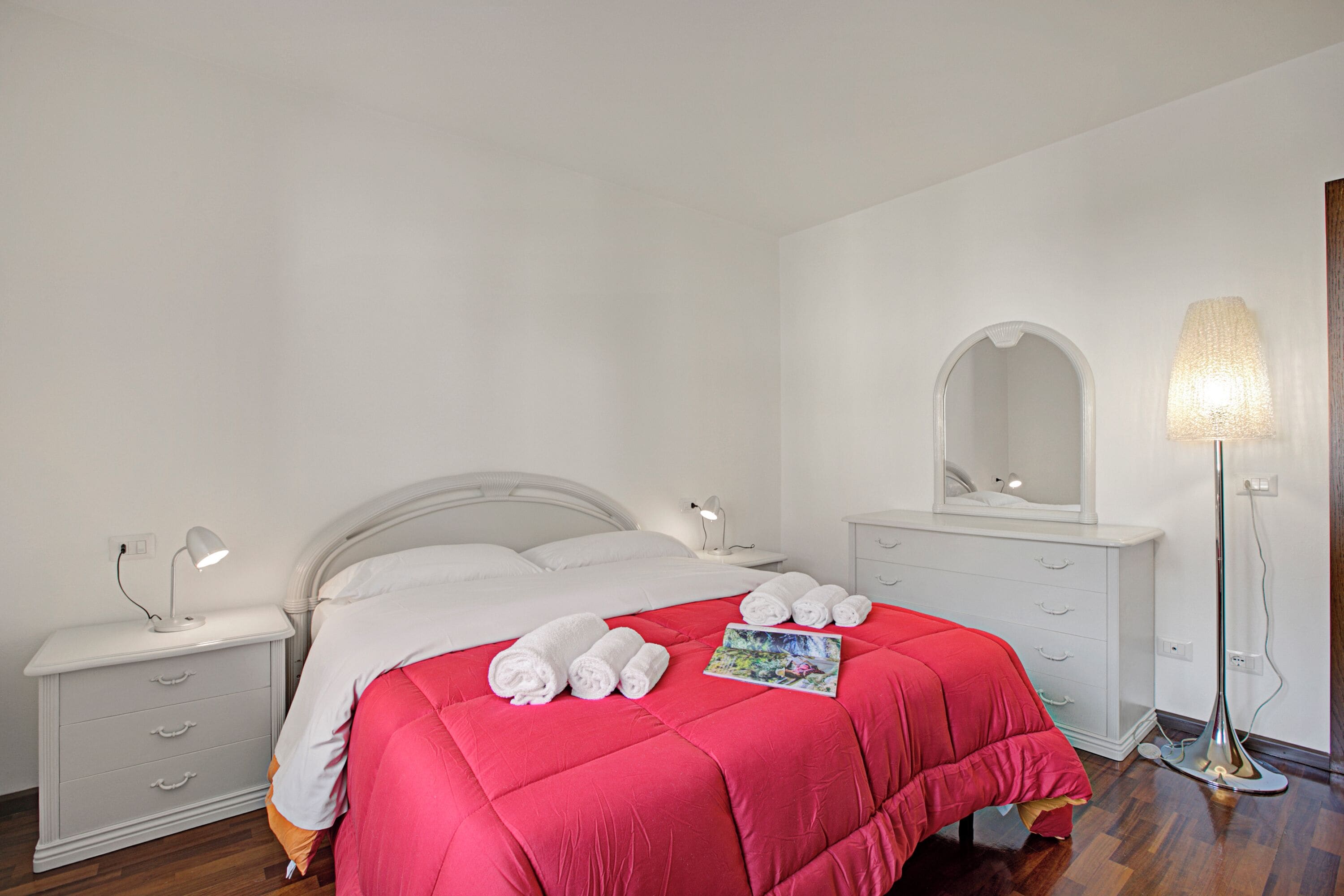 A neatly made bed with a red blanket, white pillows, and rolled towels. There is a bedside table with a lamp, a dresser with drawers, and a floor lamp.
