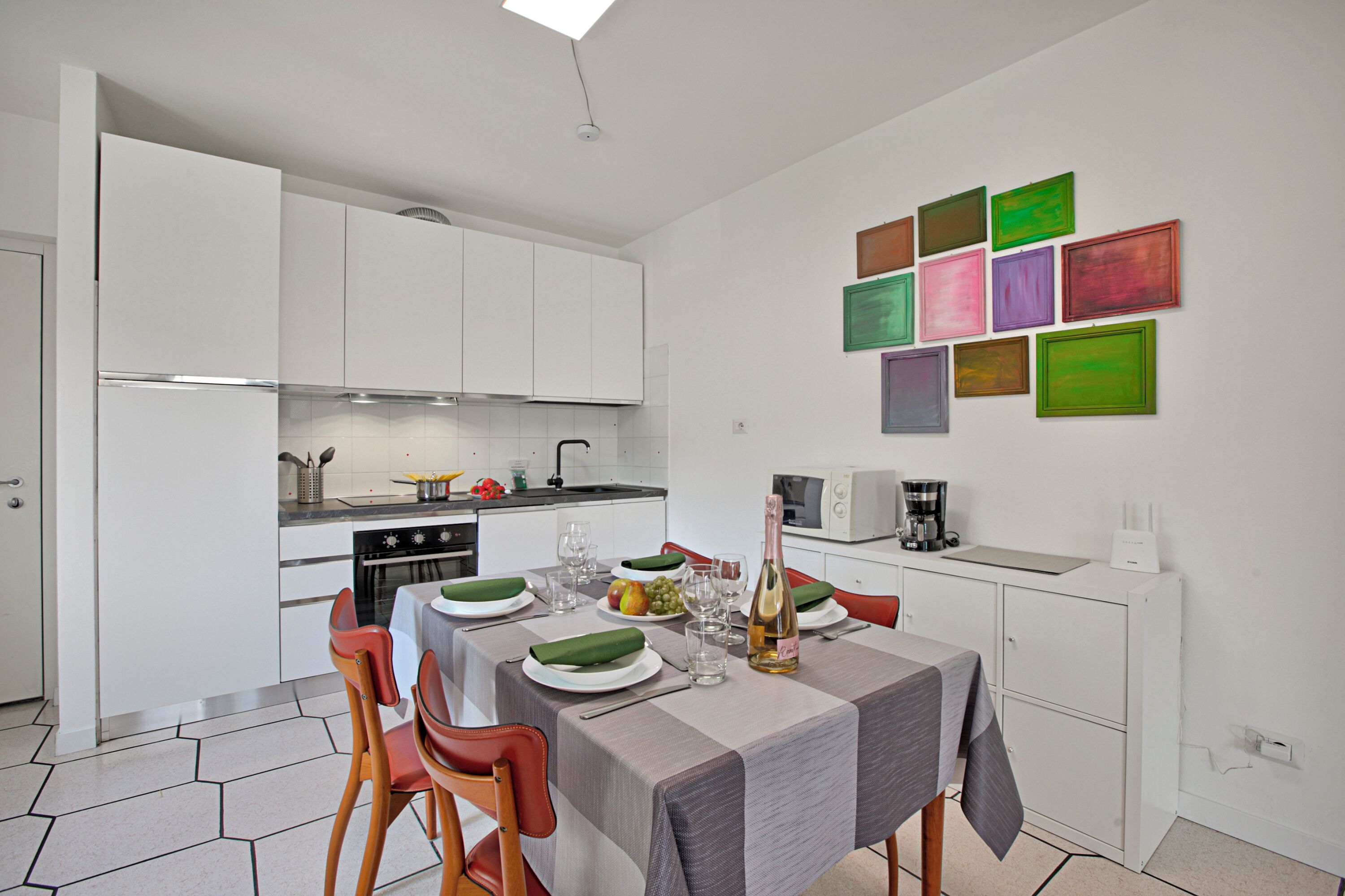 A modern kitchen with white cabinets, a dining table set with plates, glasses, and a bottle, and a wall with colorful framed pictures.