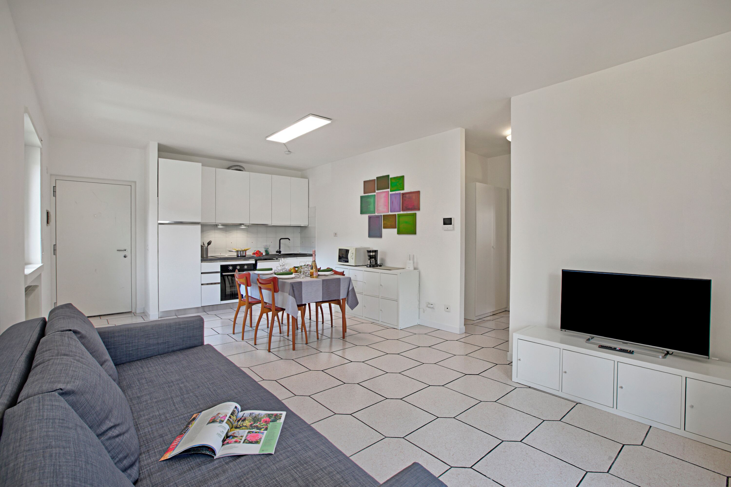 A modern living room with a grey sofa, a dining area with a table and chairs, a kitchen in the background, and a flat-screen TV on a white cabinet.
