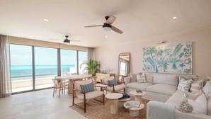 Amplia sala de estar con un ventanal que da al océano, ventilador de techo, mesa de comedor con sillas y sofá con cojines decorativos.