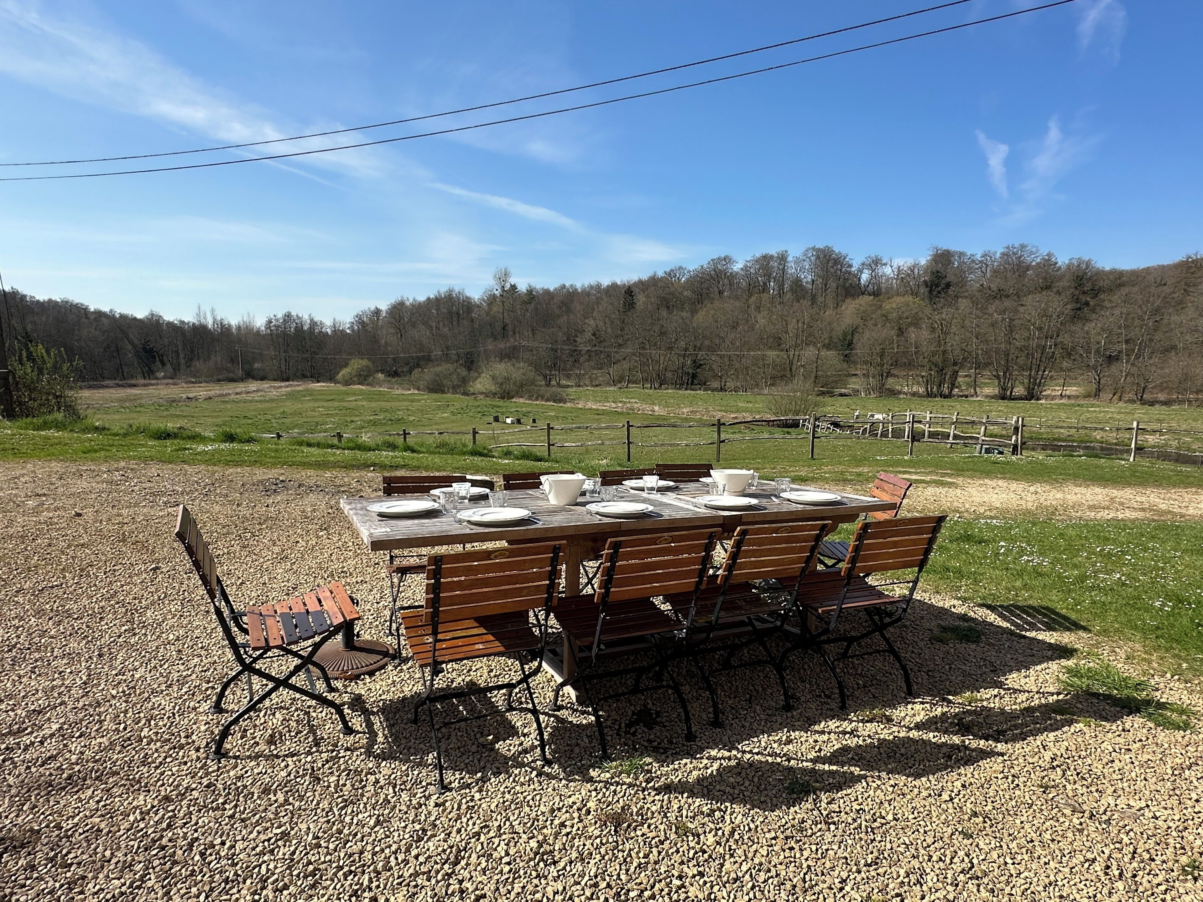 A wooden table set outdoors with chairs, surrounded by gravel, with a fence and trees in the background.