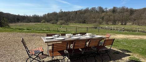 A wooden table set outdoors with chairs, surrounded by gravel, with a fence and trees in the background.