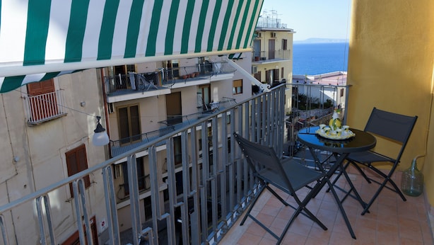 Ein Balkon mit einem grün-weiß gestreiften Jalousievorhang, einem Tisch mit Stühlen und Blick aufs Meer.