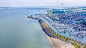Eine Luftaufnahme eines Küstenhafens mit vertäuten Booten, einem Strand mit Menschen und einer Brücke im Hintergrund.