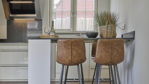 Una cucina moderna con isola centrale, due sgabelli da bar, un'ampia finestra con tende e una pianta in vaso.