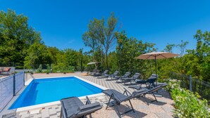 A rectangular swimming pool with a tiled deck, surrounded by lounge chairs and umbrellas, set against a backdrop of trees and a clear sky.