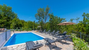 Una piscina rectangular con solárium empedrado, rodeada de sillas de descanso y sombrillas, con árboles y un cielo despejado de fondo.