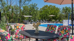 Un área junto a la piscina con una mesa redonda, sillas coloridas y sillones de descanso bajo sombrillas.