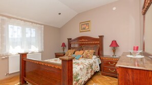 A bedroom with a wooden bed, two bedside tables, a nightstand, a radiator, a window with sheer curtains, and a framed picture on the wall.