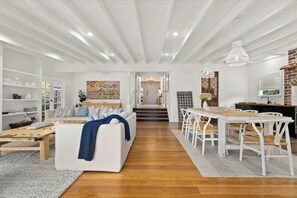 A spacious living room with a white sofa, wooden coffee table, and a dining area with white chairs and a wooden table. - Conifer - Pet Friendly with Outdoor Spa (Callala Beach)
