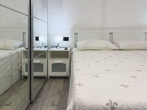 Habitación de hotel con armario espejo, mesita de noche con lámpara y cama con cubrecama estampado de flores.