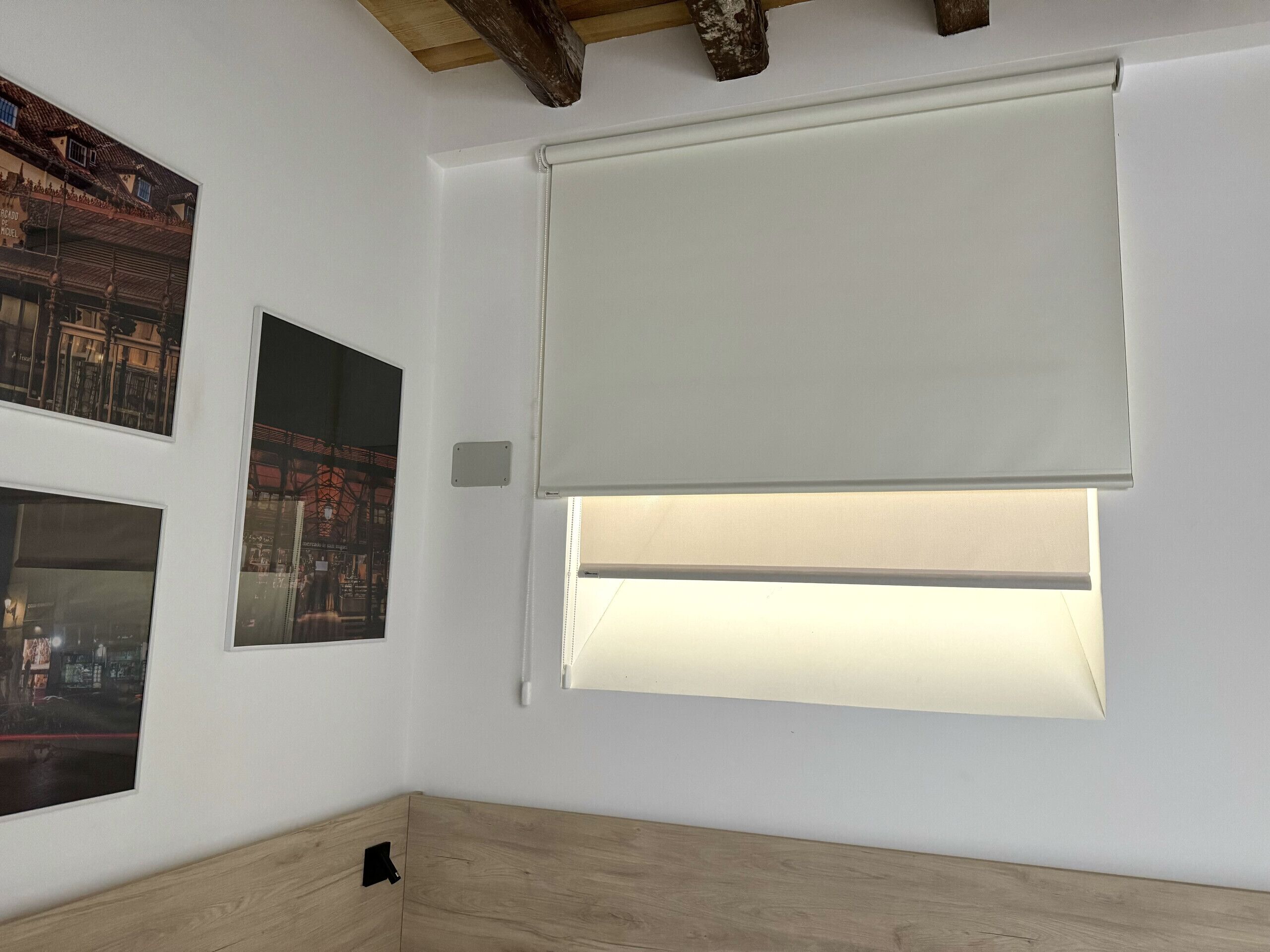 A room with a large window covered by a white roller blind, wooden flooring, and two framed pictures on the wall.