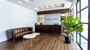 A reception area with a brown leather sofa, a white round table, a large potted plant, and a reception desk with a patterned backdrop. - Earnest Hotel Georgetown (George Town)