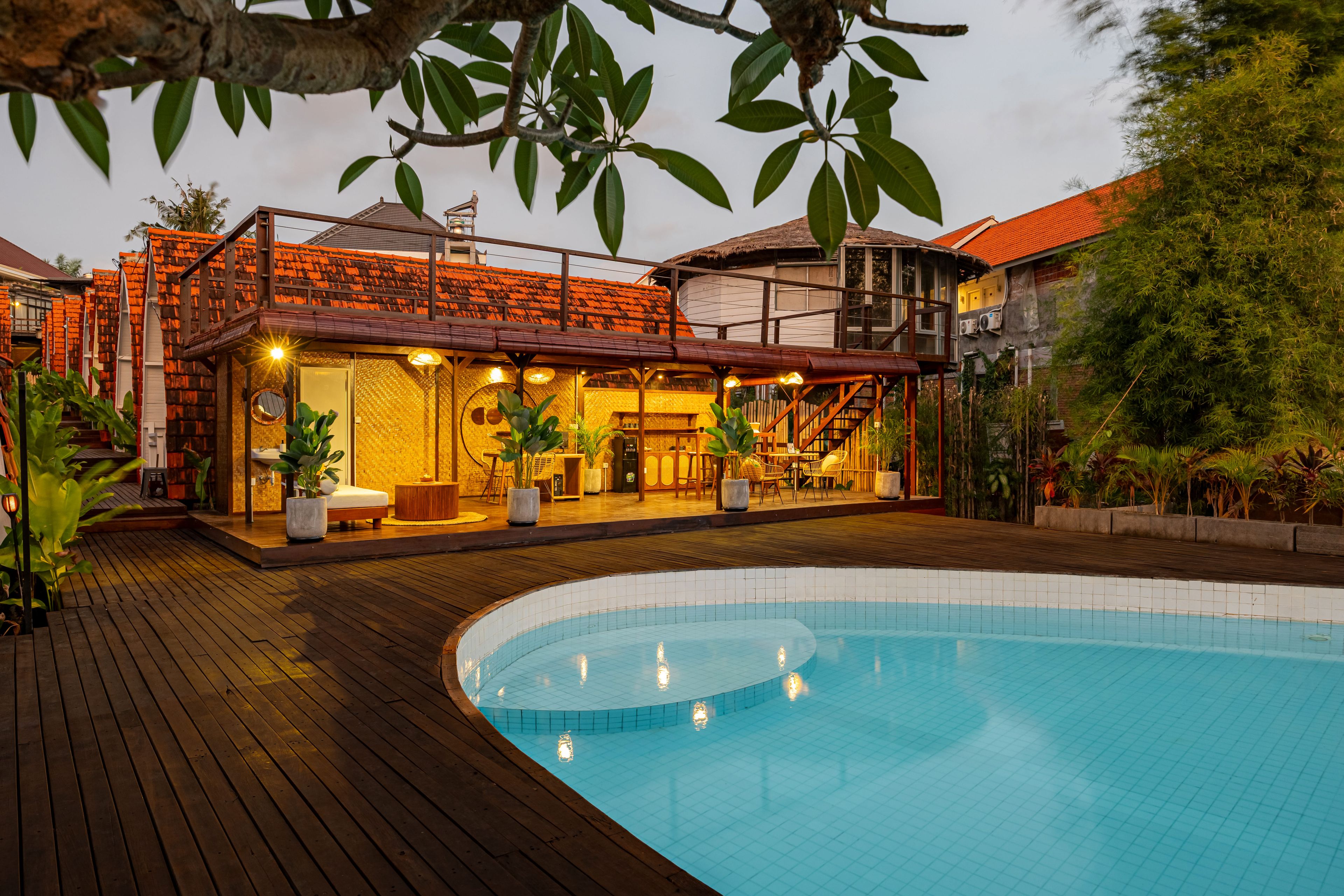 A poolside area with a wooden deck, a pool, and a building with a tiled roof and outdoor seating.