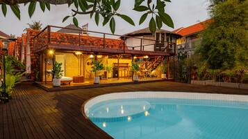 A poolside area with a wooden deck, a pool, and a building with a tiled roof and outdoor seating.