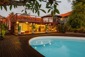 Un área junto a la piscina con muelle de madera, piscina y edificio con tejado de azulejos y asientos al aire libre.