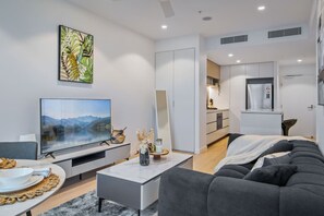 Ein modernes Wohnzimmer mit einem Flachbildfernseher, einem grauen Sofa und einem Essbereich mit Tisch für zwei Personen. Im Hintergrund ist eine Küche mit weißen Schränken und einem Kühlschrank zu sehen.