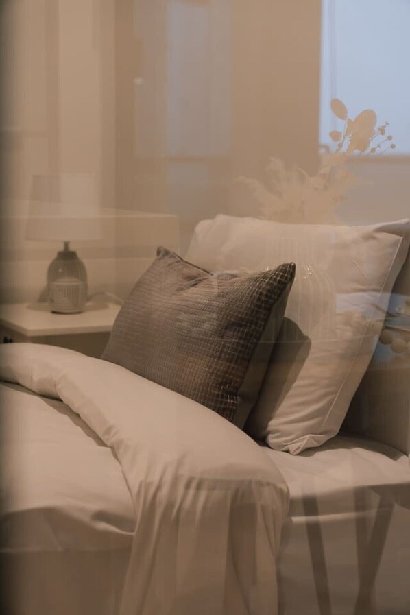 Una cama con ropa de cama blanca y un almohadón texturizado, una mesita de noche con una lámpara y una ventana con vista al cielo.