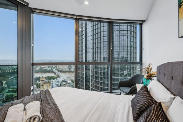 A modern bedroom with a large window offering a city view, a bed with pillows, a chair, and a vase with flowers.