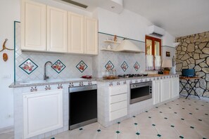 Una cocina con armarios blancos, estufa, horno y lavamanos. Se aprecia una pared de piedra y una ventana con cortinas.
