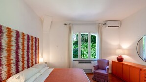 Un dormitorio con cama, una silla, una ventana con cortinas y una lámpara sobre un mueble.
