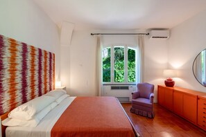 Una camera da letto con un letto, una sedia, una finestra con tende e una lampada su un mobiletto.