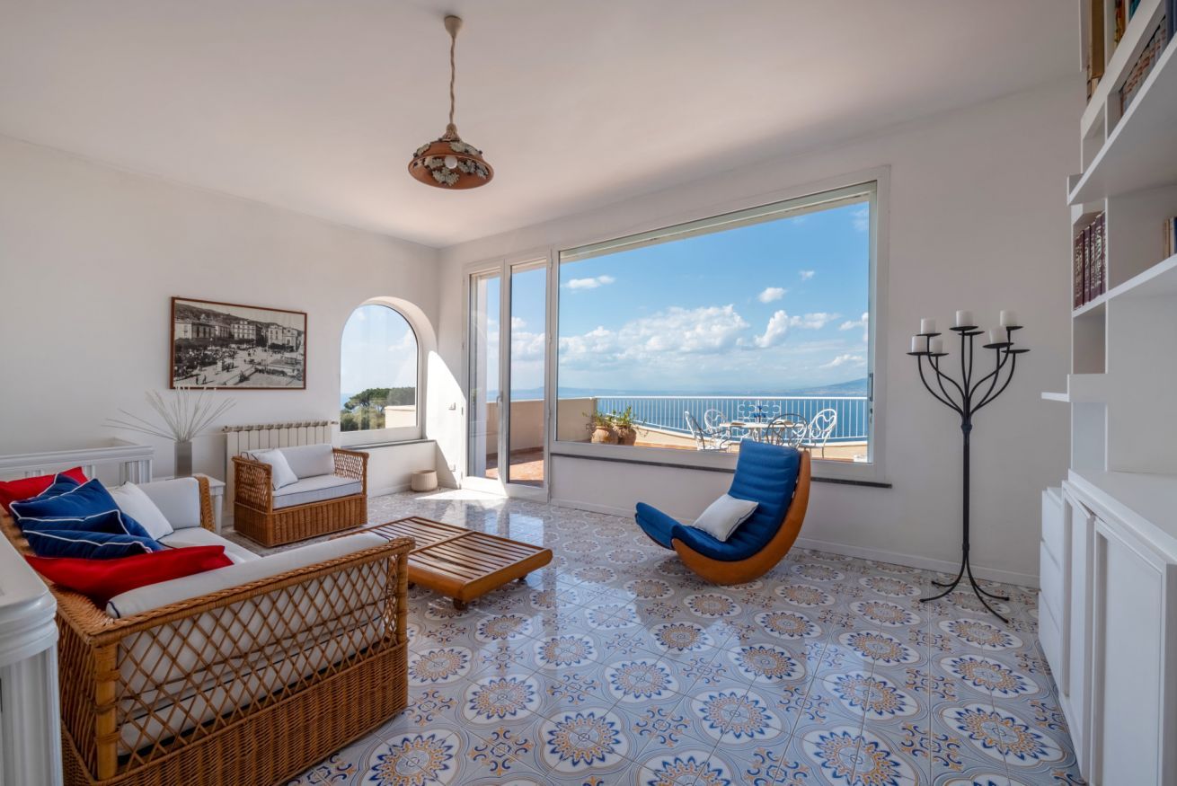 Uma sala de estar espaçosa com uma grande janela que oferece vista para o mar, um sofá de vime, uma poltrona azul e um chão com padrão.