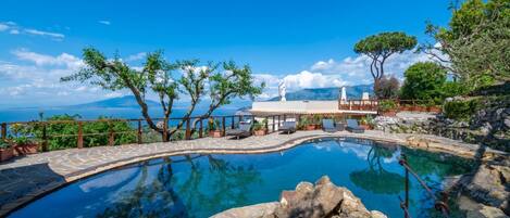 A clear blue pool with a stone deck, surrounded by trees and a view of the sea.