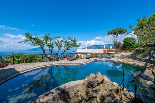 A clear blue pool with a stone deck, surrounded by trees and a view of the sea.