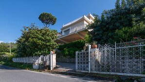 Una casa de dos pisos con balcón, rodeada por una cerca blanca y abundante vegetación.