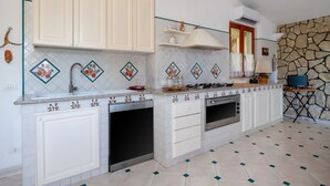 Una cocina con armarios blancos, estufa, horno y lavamanos. Se aprecia una pared de piedra y una ventana con cortinas.