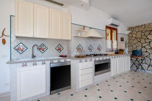 Cucina con mobili bianchi, piano cottura, forno e lavello. Si nota una parete in pietra e una finestra con tende.