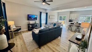 A modern living room with a dark sofa, a TV, and a dining area with a kitchen in the background. - BRAND NEW 3 Bedroom, 2.5 Bath House (High Point)
