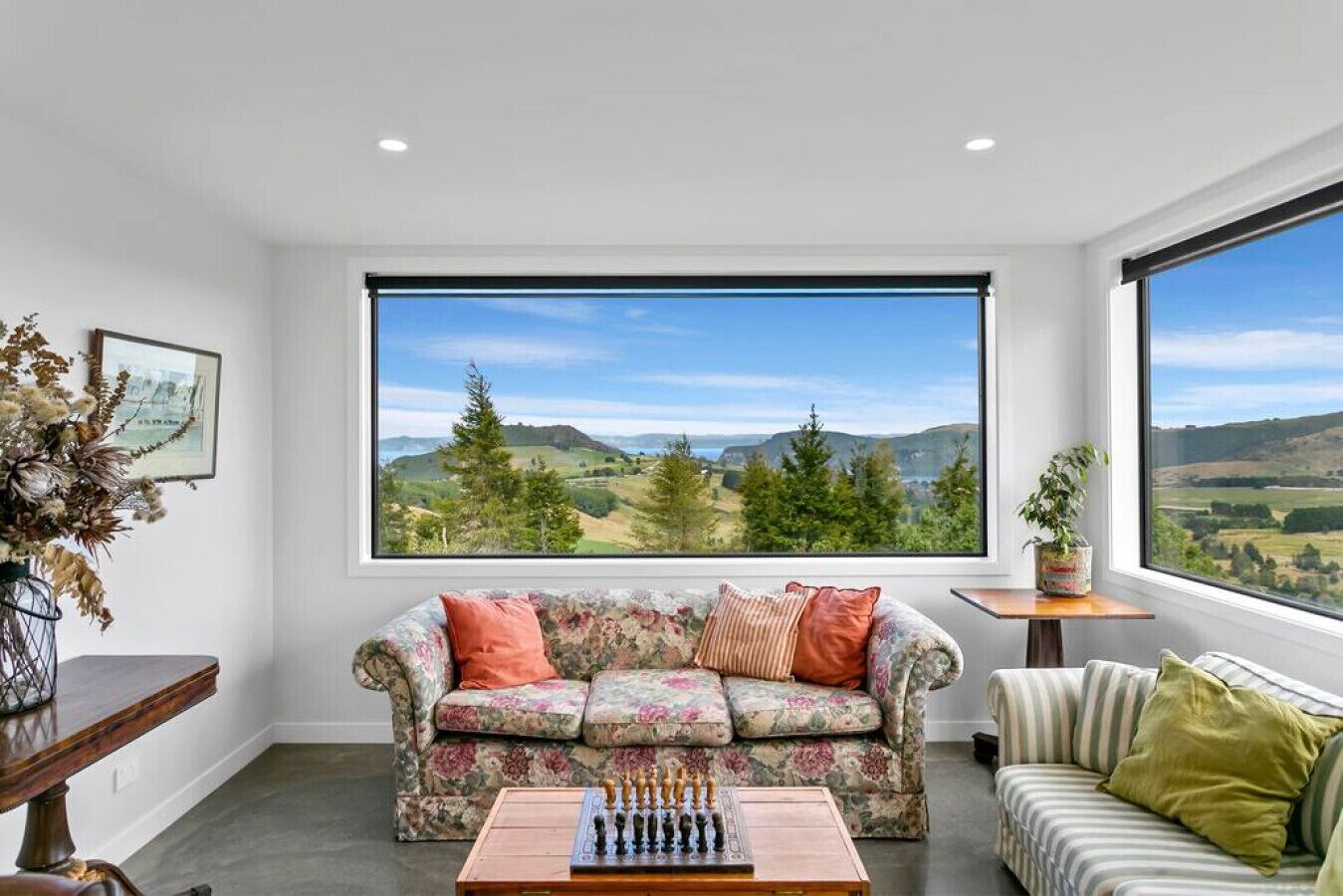 A living room with a large window, a floral sofa, a chess table, and a view of rolling hills.