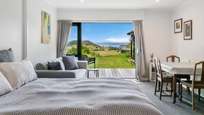 A bedroom with a large bed, a sofa, a dining table with chairs, and a view of a green landscape through a large window. - Peaceful Luxury Retreat - with Spa Pool, a spacious, beautiful, private property with stunning views (Taupō)