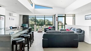 Uma sala de estar moderna com um sofá preto grande, uma área de jantar com banquetas e grandes janelas com vista para o exterior.