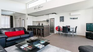 Uma sala de estar moderna com um sofá de couro preto, uma mesa de centro de vidro e uma almofada vermelha. Há uma área de cozinha com balcão e banquetas, e uma mesa de jantar com cadeiras. Uma TV de tela plana está fixada na parede.